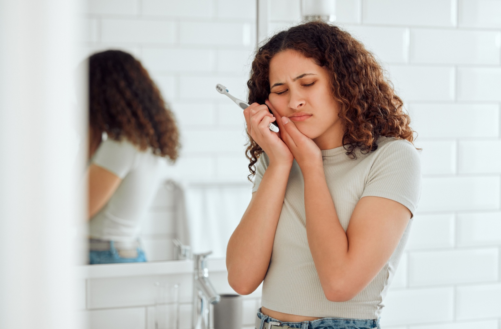 A person in the bathroom holding the side of their face in pain from a tooth ache holding a toothbrush.