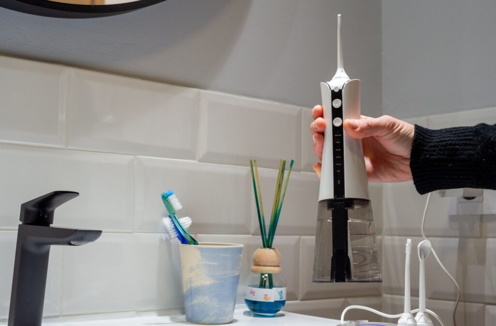A hand holding a water flosser next to a cup of toothbrushes in a bathroom.