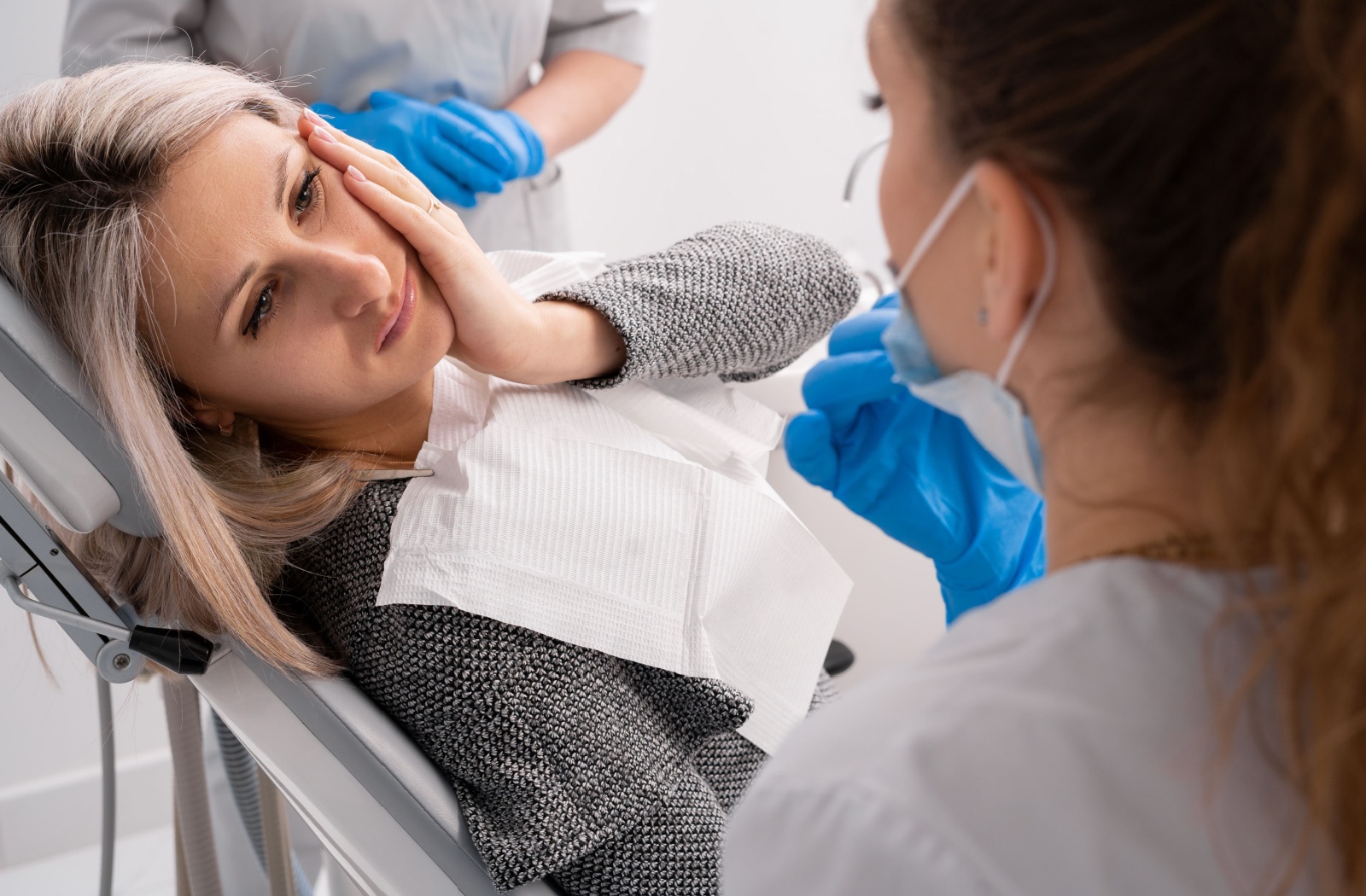A person laying down in a dental chair and holding their jaw due to discomfort caused by a dental emergency